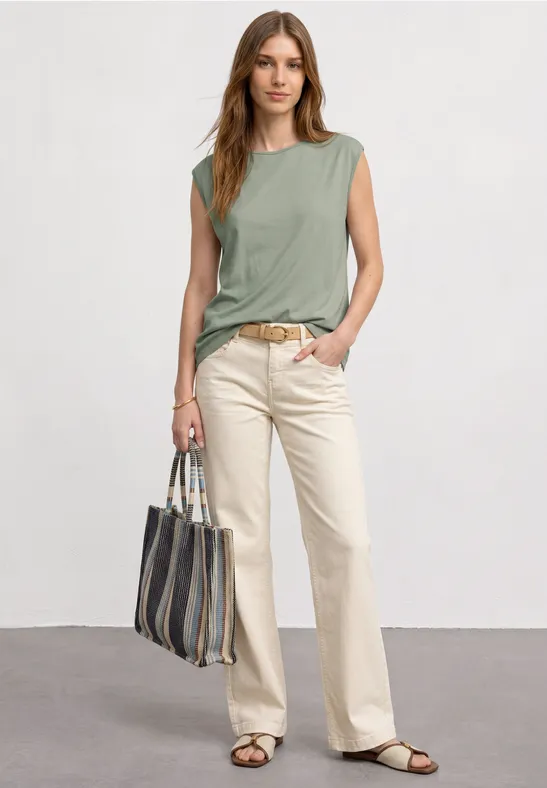 Een vrouw in een groen mouwloos shirt, lichtgekleurde broek, sandalen en een gestreepte tas.