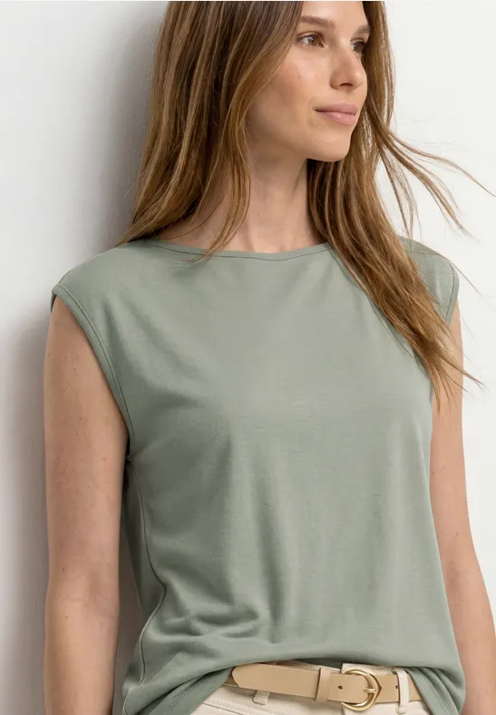 Een vrouw draagt een mouwloos olijfgroen topje met een beige riem tegen een lichte muur.
