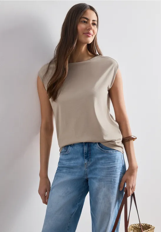 Frau in ärmellosem beigefarbenem Top und blauen Jeans hält eine braune Tasche.