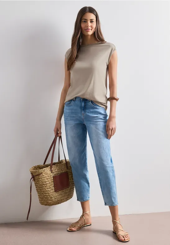 Frau in beigem Top, blauen Jeans, Sandalen und großer Korbtasche vor neutralem Hintergrund.