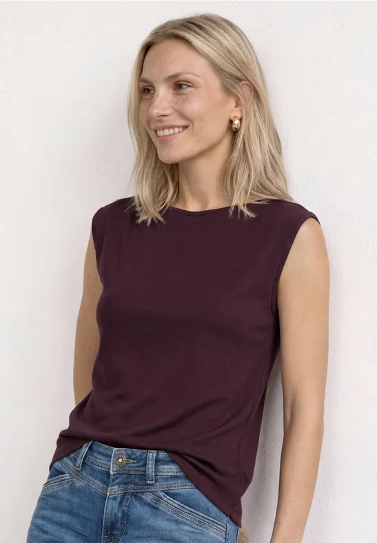 Vrouw in bordeauxkleurige mouwloze top en blauwe jeans, leunend tegen een witte muur.