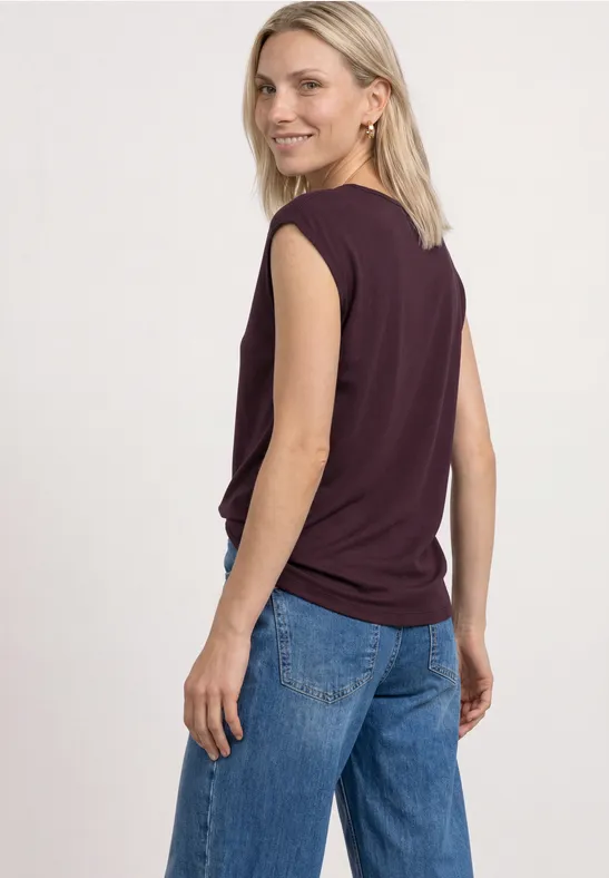 Een persoon met blond haar draagt een mouwloos donkerpaars topje en een blauwe spijkerbroek, kijkt achterom.