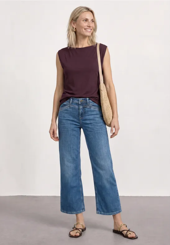 Vrouw voor neutrale achtergrond in paars topje, wijde jeans, zwarte sandalen en rieten tas.