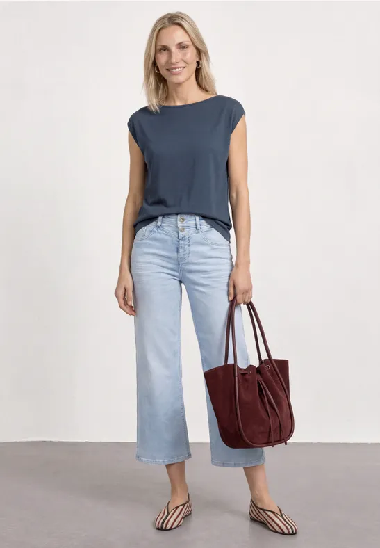 Vrouw met marineblauw mouwloos topje, lichtblauwe wijde jeans, bruine sandalen en bordeaux suède tas.