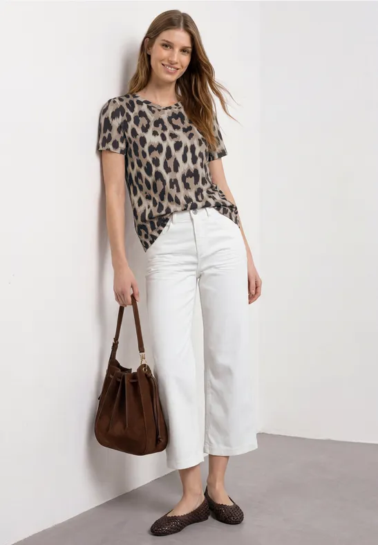 Vrouw leunt tegen witte muur, luipaardshirt, witte culotte, bruin tasje en schoenen.