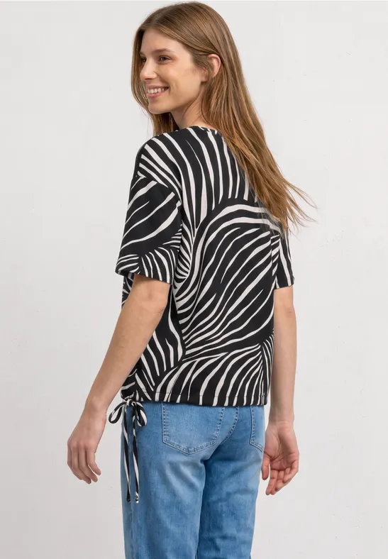 Femme portant un top noir à imprimé zèbre blanc, manches mi-longues et cordons sur le côté, avec jean bleu.