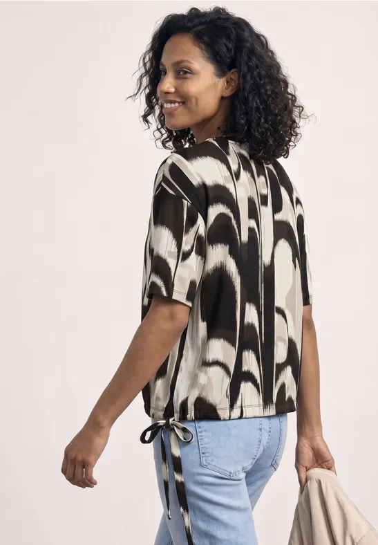 Frau mit lockigen Haaren im schwarz-weißen Shirt mit seitlichen Schleifen, Jeans und Schultertasche.