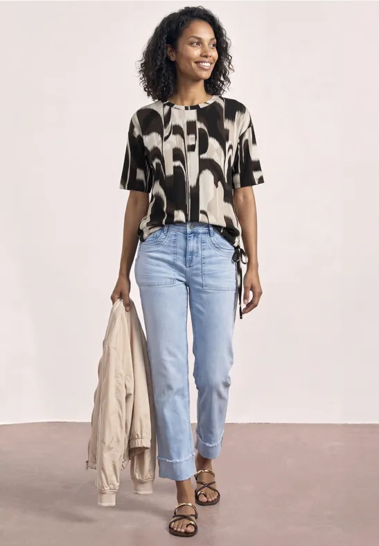 Frau in gemusterter Bluse, hellblauen Jeans und Sandalen hält eine Jacke in der Hand.