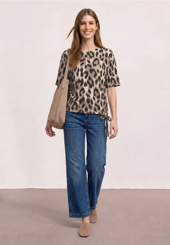 Eine Frau mit Leopard-Shirt, blauen Jeans, beigen Loafers und beiger Tasche vor hellem, neutralem Hintergrund.