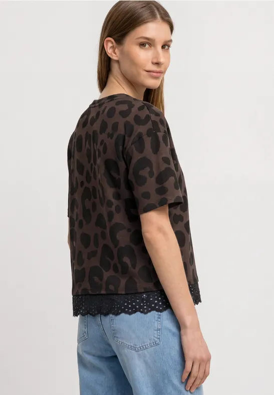 Eine Frau steht im Profil und trägt eine braune Leo-Tunika mit kurzen Ärmeln und hellblaue Jeans.