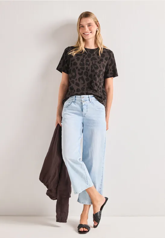Frau in gemustertem Shirt, hellen Jeans und schwarzen Sandalen lehnt entspannt an der Wand, Jacke in der Hand.