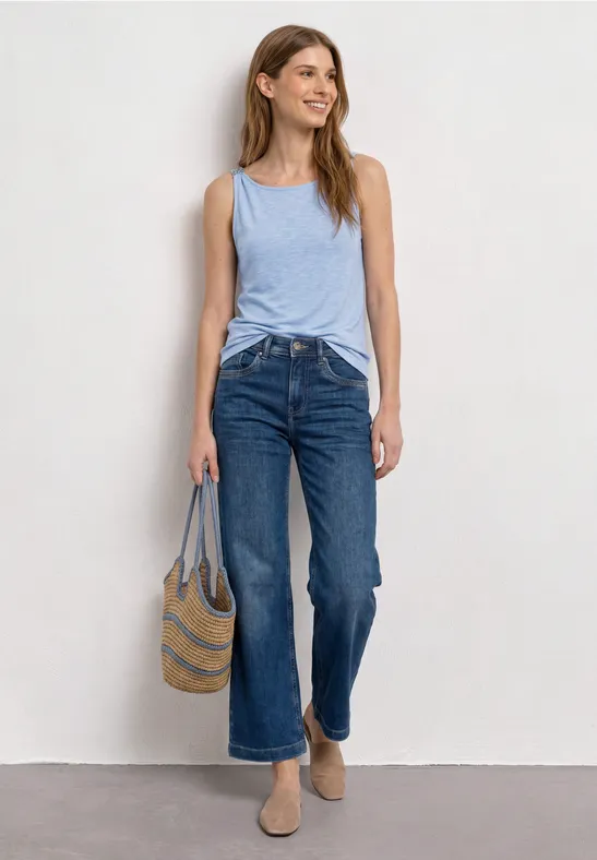 Frau in hellblauem Tanktop, weitem Jeans, mit geflochtener Tasche und beigen Slippers