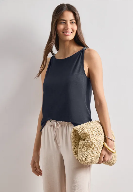 Eine Frau trägt ein ärmelloses schwarzes Top, bequeme helle Hosen und hält eine Strohtasche.