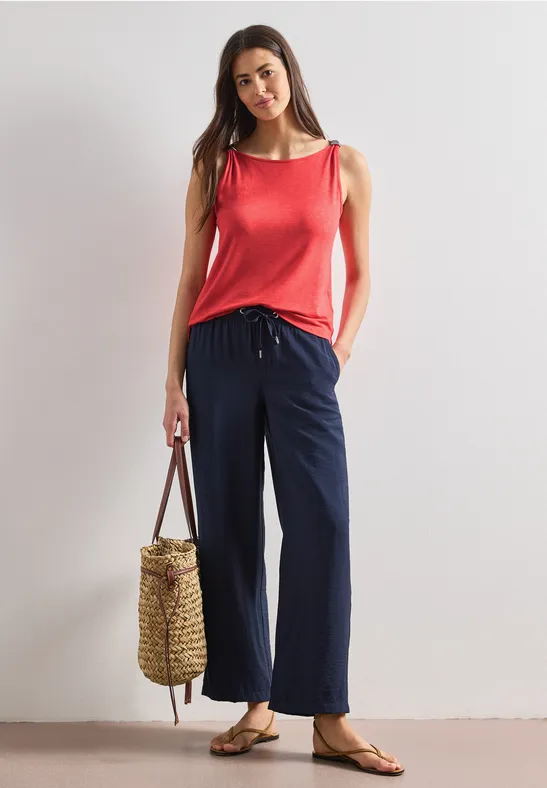 Frau in rotem Top, weiter blauer Hose, Sandalen und Korbtasche vor neutralem Hintergrund.