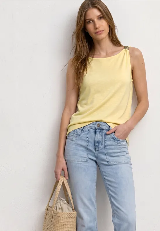Frau vor neutralem Hintergrund trägt gelbes Top, blaue Jeans und hält Tasche.