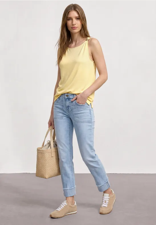Frau trägt gelbes Top, blaue Jeans, beige Sneaker und Strohtasche vor neutralem Hintergrund.
