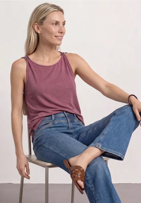 Frau sitzt auf Stuhl, trägt rosafarbenes Tanktop, blaue Jeans und braune Loafers.