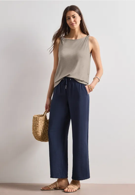 Frau in ärmellosem Top und weiten blauen Hosen, mit Strohtasche und Sandalen.