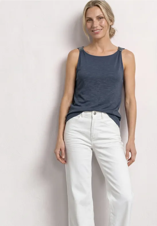 Frau in blauem ärmellosen Top und weißen Jeans steht vor neutralem Hintergrund.