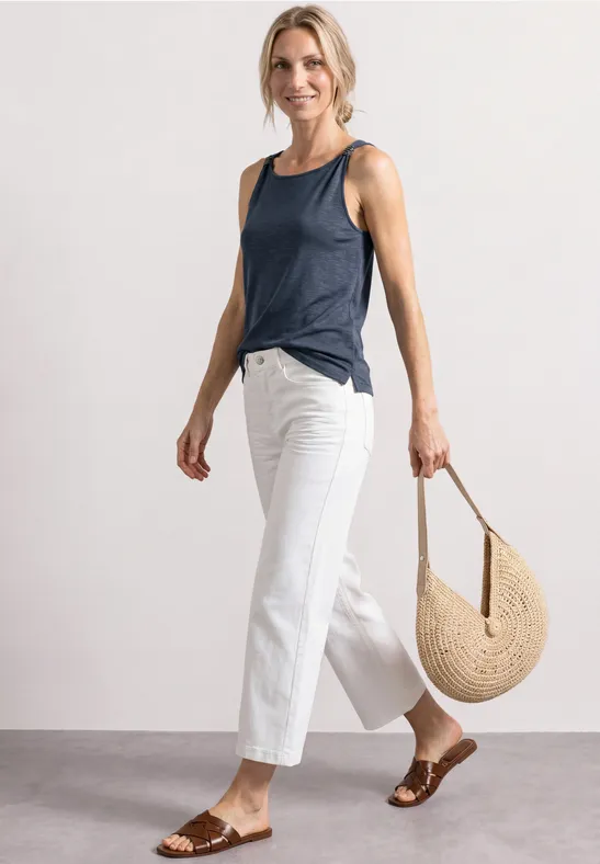 Frau in weißer, weiter Hose, blauem ärmellosem Top, braunen Sandalen und runder Basttasche.