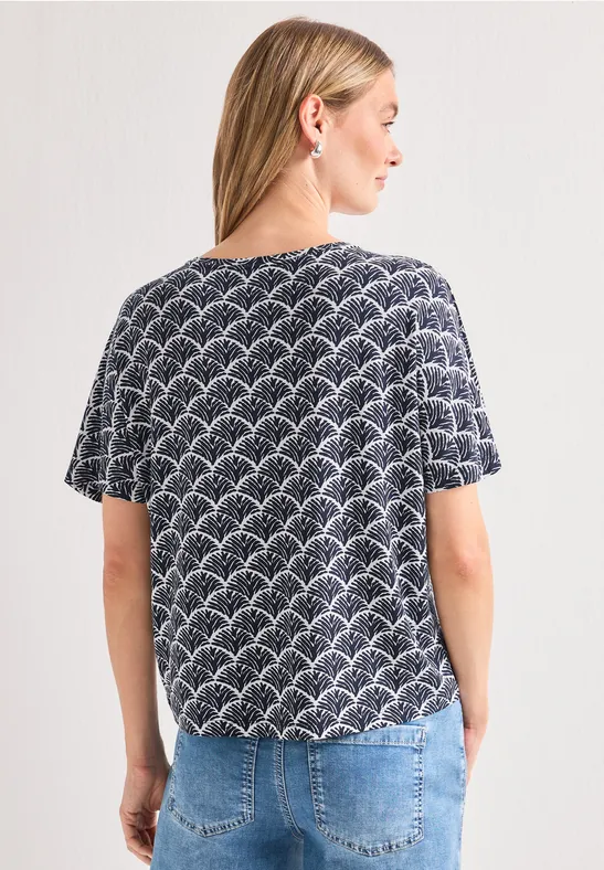 Frau von hinten in T-Shirt mit schwarz-weißem Bogenmuster, blauer Jeans und Zopf.