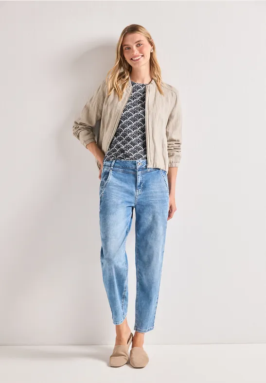 Frau in beiger Jacke, gemustertem T-Shirt, hellblauen Jeans und beigen Slippern vor neutralem Hintergrund.