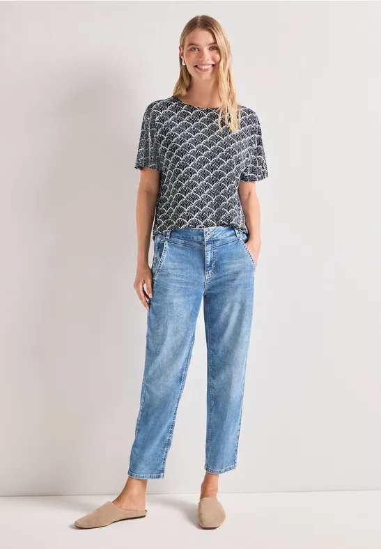 Frau mit grauem Muster-Shirt, blauen Jeans und beigen Pantoletten vor hellem Hintergrund.