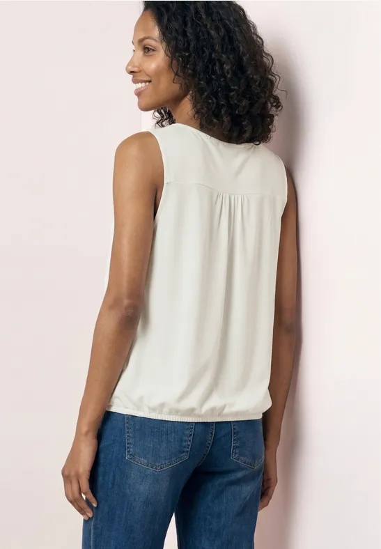 Vrouw van achteren in een witte mouwloze top en blauwe spijkerbroek voor een lichtroze muur.