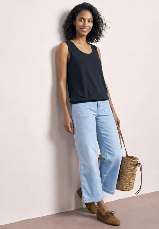 Vrouw leunt tegen een lichte muur, zwart topje, wijde lichte jeans, bruine loafers en een gevlochten mandtas.