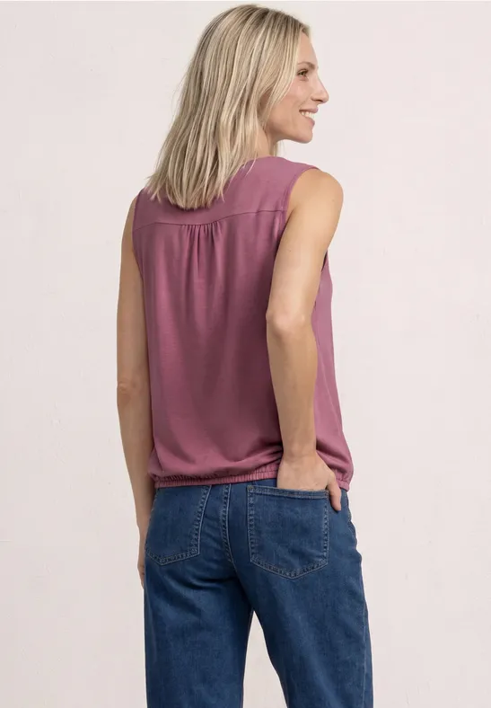 Vrouw van achteren in een mouwloze roze top en blauwe spijkerbroek, één hand in de zak.