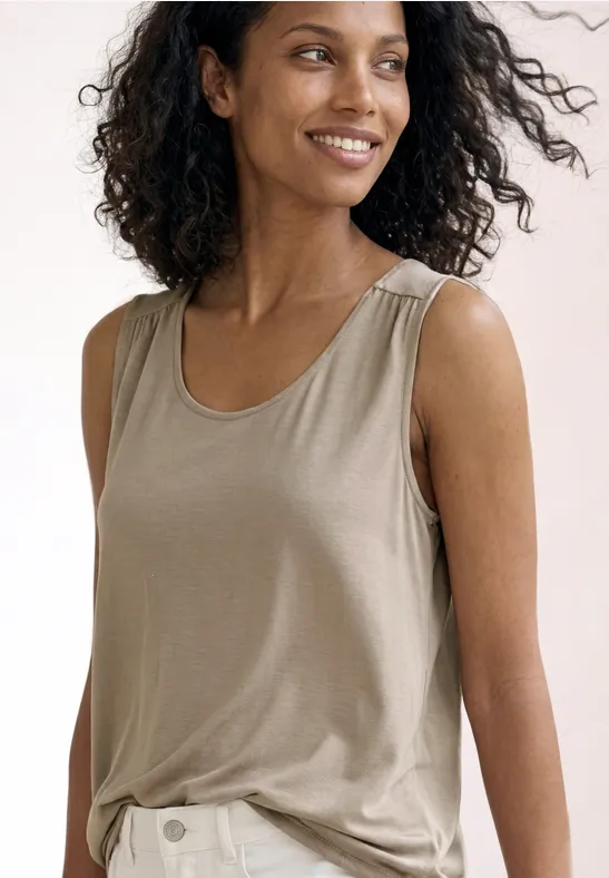 Femme aux cheveux bouclés portant un débardeur beige sur fond clair.