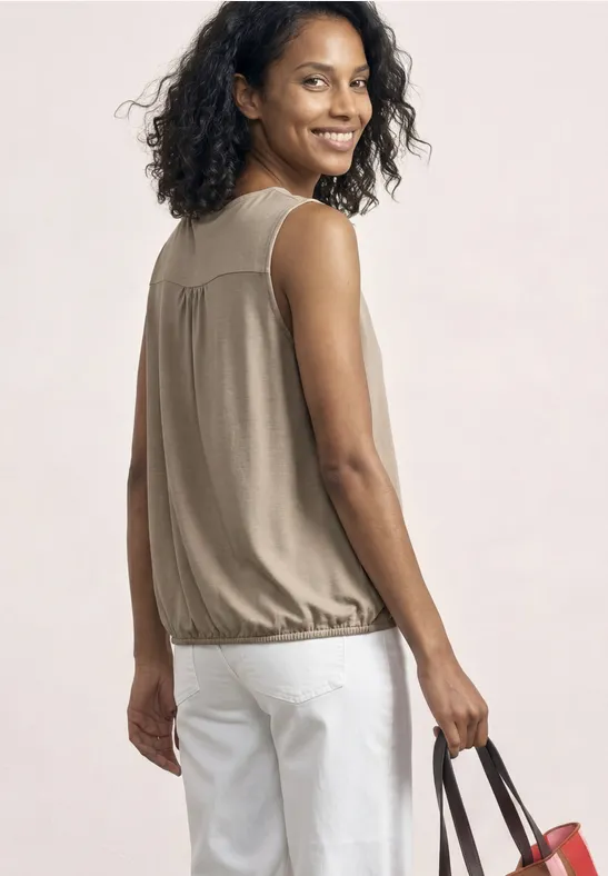 Femme vue de dos portant un débardeur beige, pantalon blanc, tenant un sac à main.