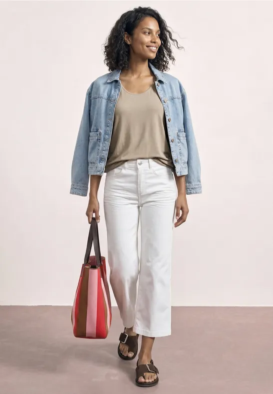 Femme en veste en denim, haut beige, pantalon blanc, sandales et sac rouge sur fond neutre.