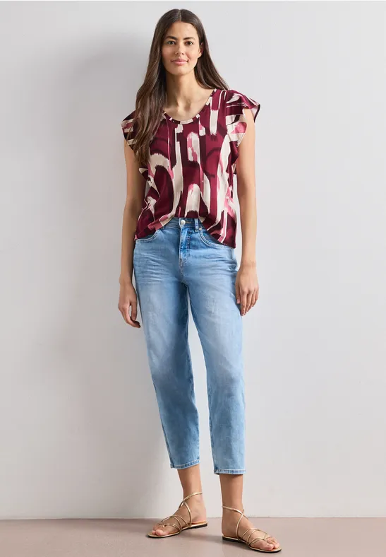 Eine Frau trägt ein kurzärmeliges, gemustertes T-Shirt, blaue Jeans und Sandalen.