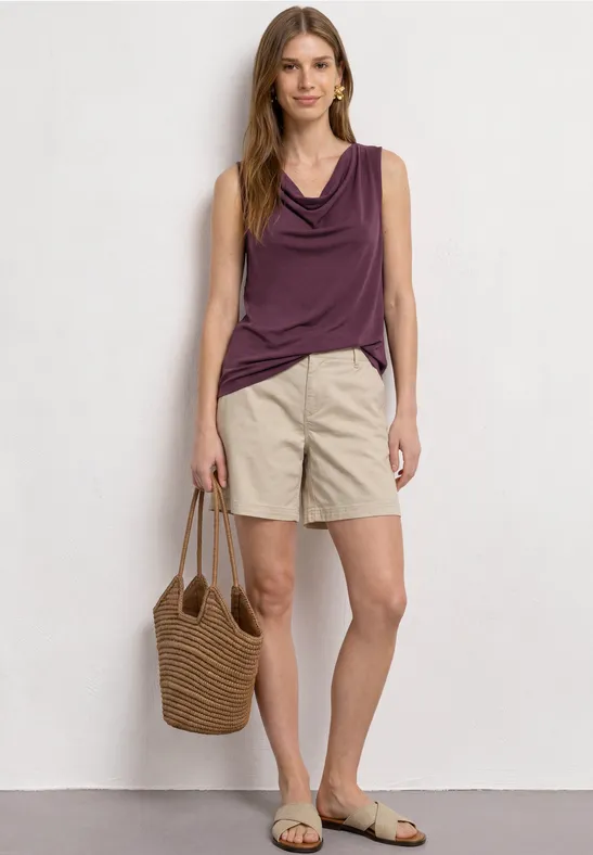 Frau trägt ärmelloses lila Top, beige Shorts, flache Sandalen und eine geflochtene Strohtasche.