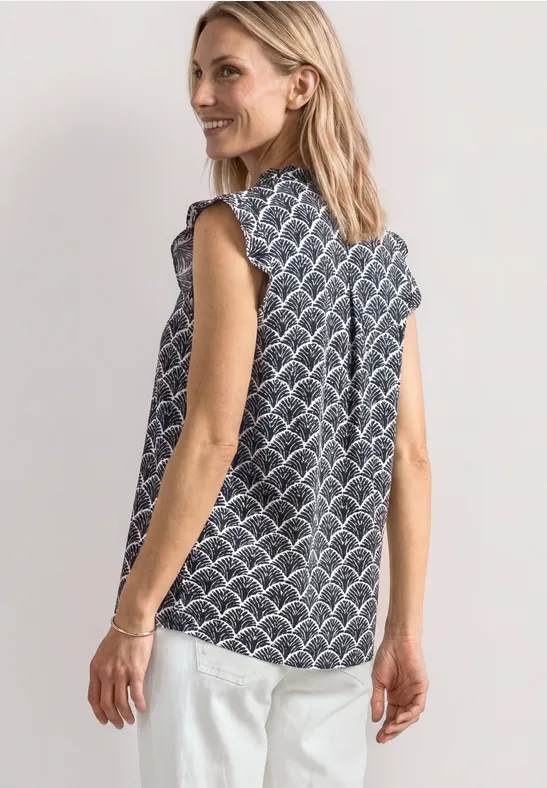 Vrouw in mouwloze blouse met patroon en witte broek, zij-aanzicht tegen een lichte achtergrond.
