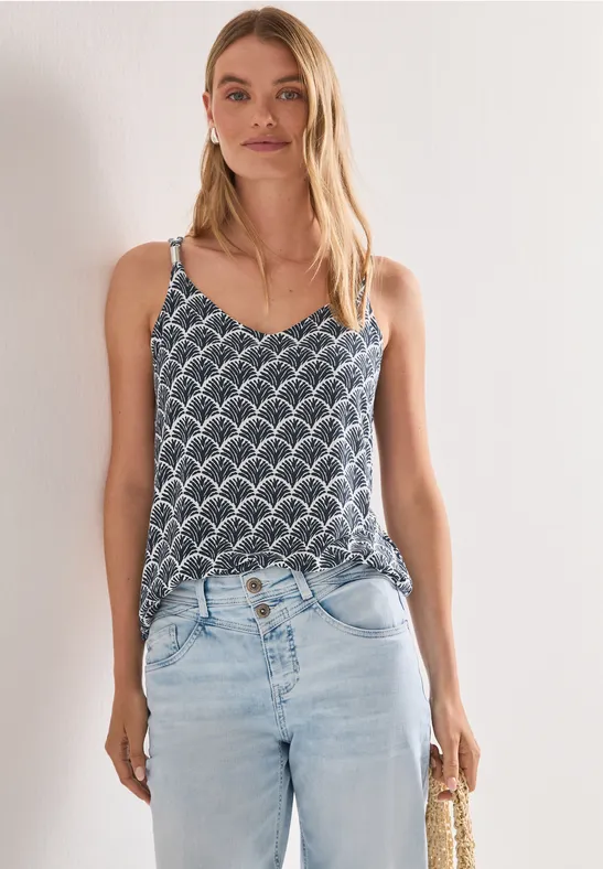 Frau trägt gemustertes Top, helle Jeans und hält eine Strohtasche vor weißem Hintergrund.