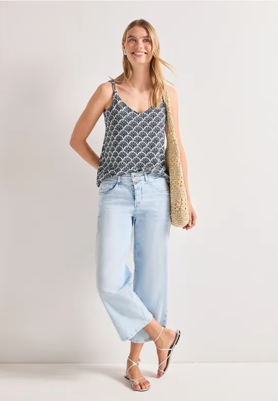Frau trägt gemustertes Top, helle Jeans, weiße Sandalen und eine Strohtasche.