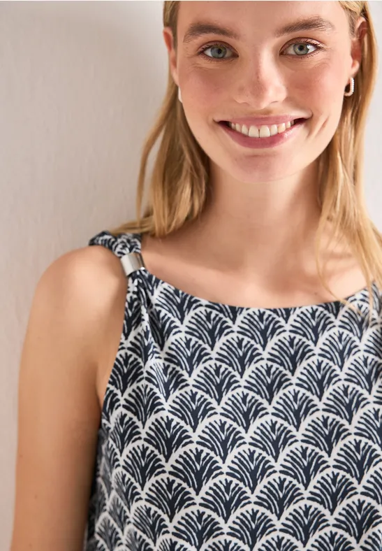 Eine junge Frau trägt ein ärmelloses Top mit schwarz-weißem Blattmuster vor beigem Hintergrund.