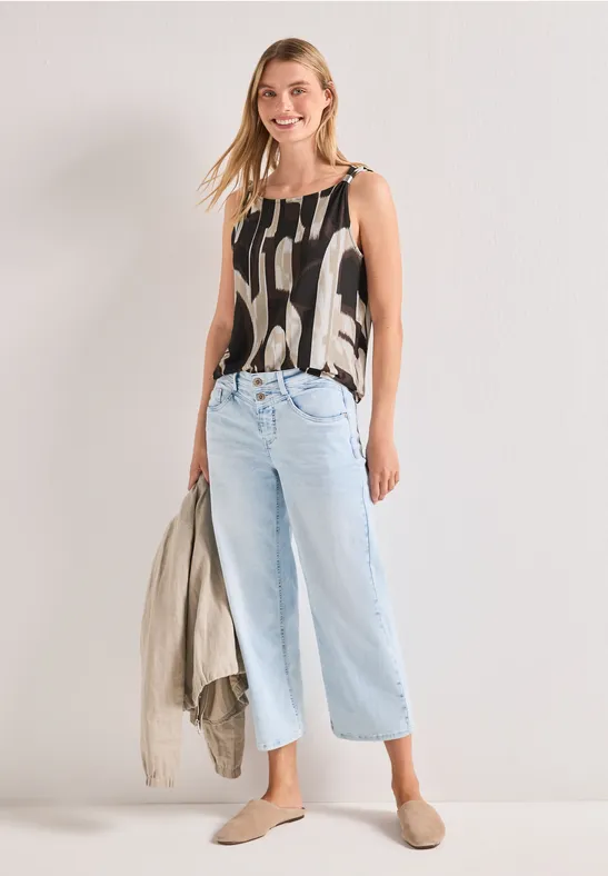 Femme portant un top imprimé, un jean large bleu clair et des mules plates, tenant une veste à la main.