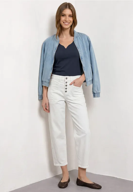 Frau mit leichtem Jeansjäckchen über den Schultern, dunkelblauem Top, weißen High-Waist-Jeans und braunen Ballerinas.
