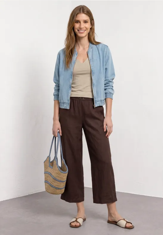 Frau in blauer Jacke, beigem Top, brauner Culotte, Sandalen und Korbtasche vor hellem Hintergrund.