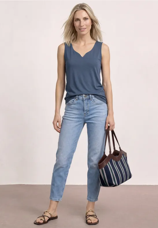 Vrouw in blauw mouwloos topje en spijkerbroek, houdend een gestreepte tas, in een ontspannen houding.