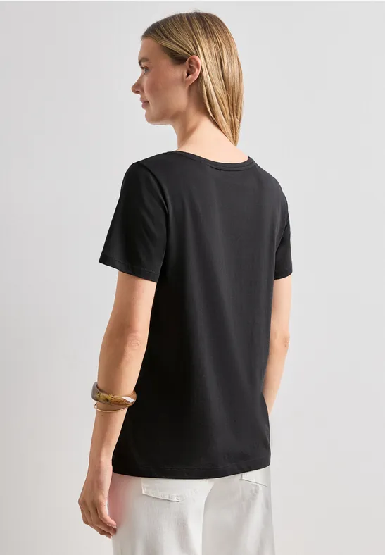 Frau von hinten in schwarzem T-Shirt und weißen Hosen, helle Haare über die Schulter fallend.