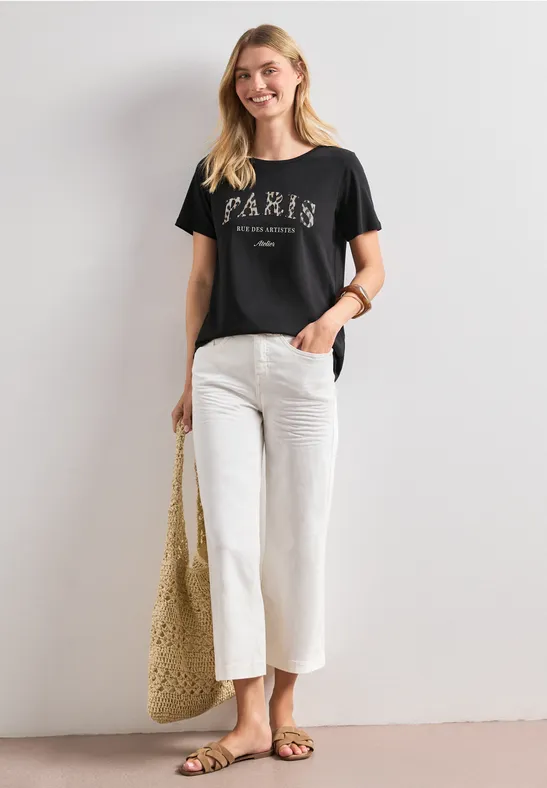 Frau in schwarzem Paris-T-Shirt, weißer Culotte, flachen Sandalen und geflochtenem Korbtasche vor weißer Wand