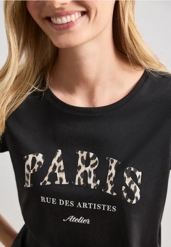 Frau in schwarzem T-Shirt mit leopardbedrucktem Schriftzug 'PARIS' und 'Rue des Artistes Atelier'.