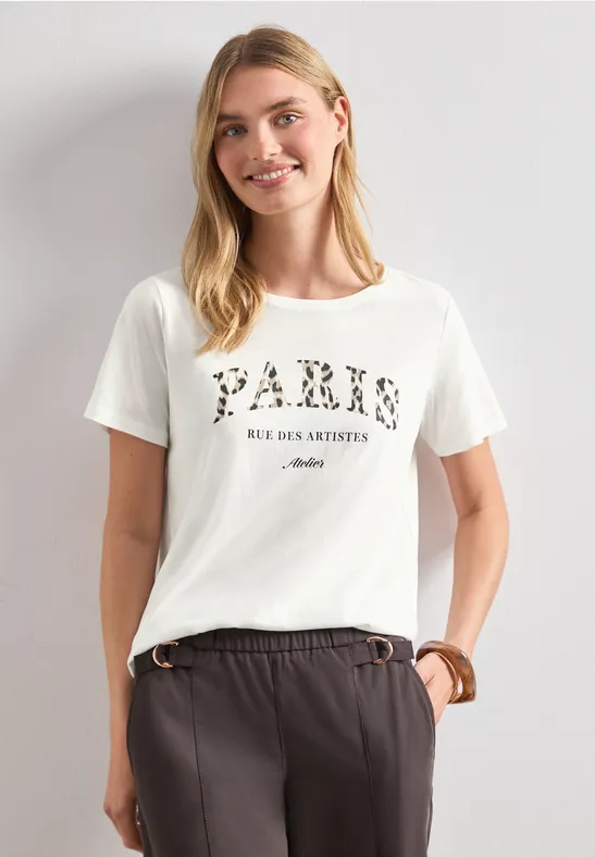 Frau vor hellem Hintergrund trägt weißes T-Shirt mit 'PARIS RUE DES ABBESSES' und dunkle Hose.