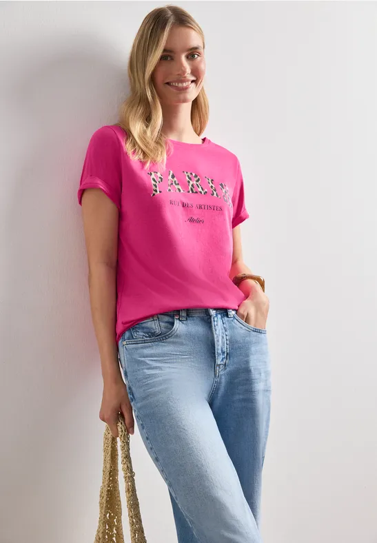 Frau in pinkem T-Shirt, hellblauen Jeans und Strohtasche steht vor weißem Hintergrund.