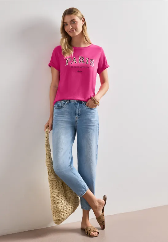Frau in pinkem T-Shirt und hellen Jeans, trägt Strohtasche und Sandalen vor weißer Wand.