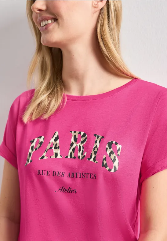 Eine Frau trägt ein pinkes T-Shirt mit Leopardenmuster-Schriftzug 'PARIS Rue des Artistes Atelier'.